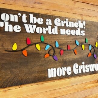 wood stained board with colored laser cut christmas lights and the words Don't be a grinch! The world needs more Griswolds laser cut from wood painted white.